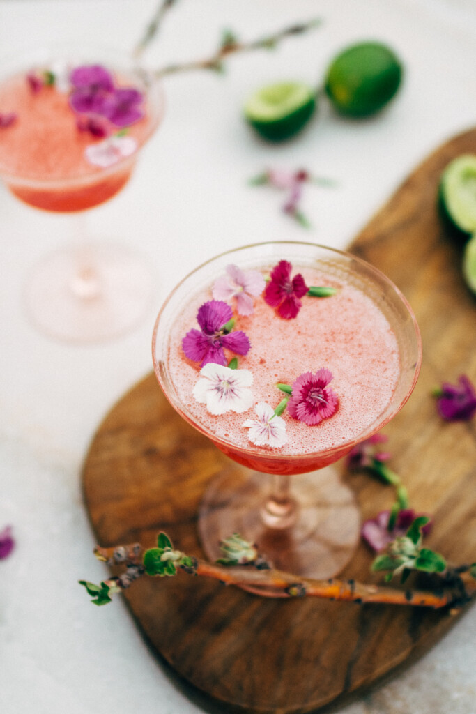 A side-view of two bright pink cocktails in pale-pink cocktail glasses, with edible purple flowers scattered on the drink's surface. There are freshly squeezed limes and scattered flowers in the background. One of the drinks is on an olive-wood cutting board.