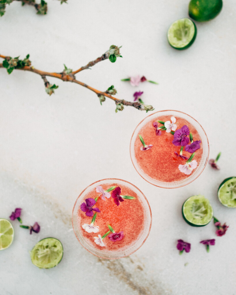 A top-down look at a bright pink cocktail with edible purple flowers scattered on the drink's surface. There are freshly squeezed limes in the background and blurred apple branches in the foreground.