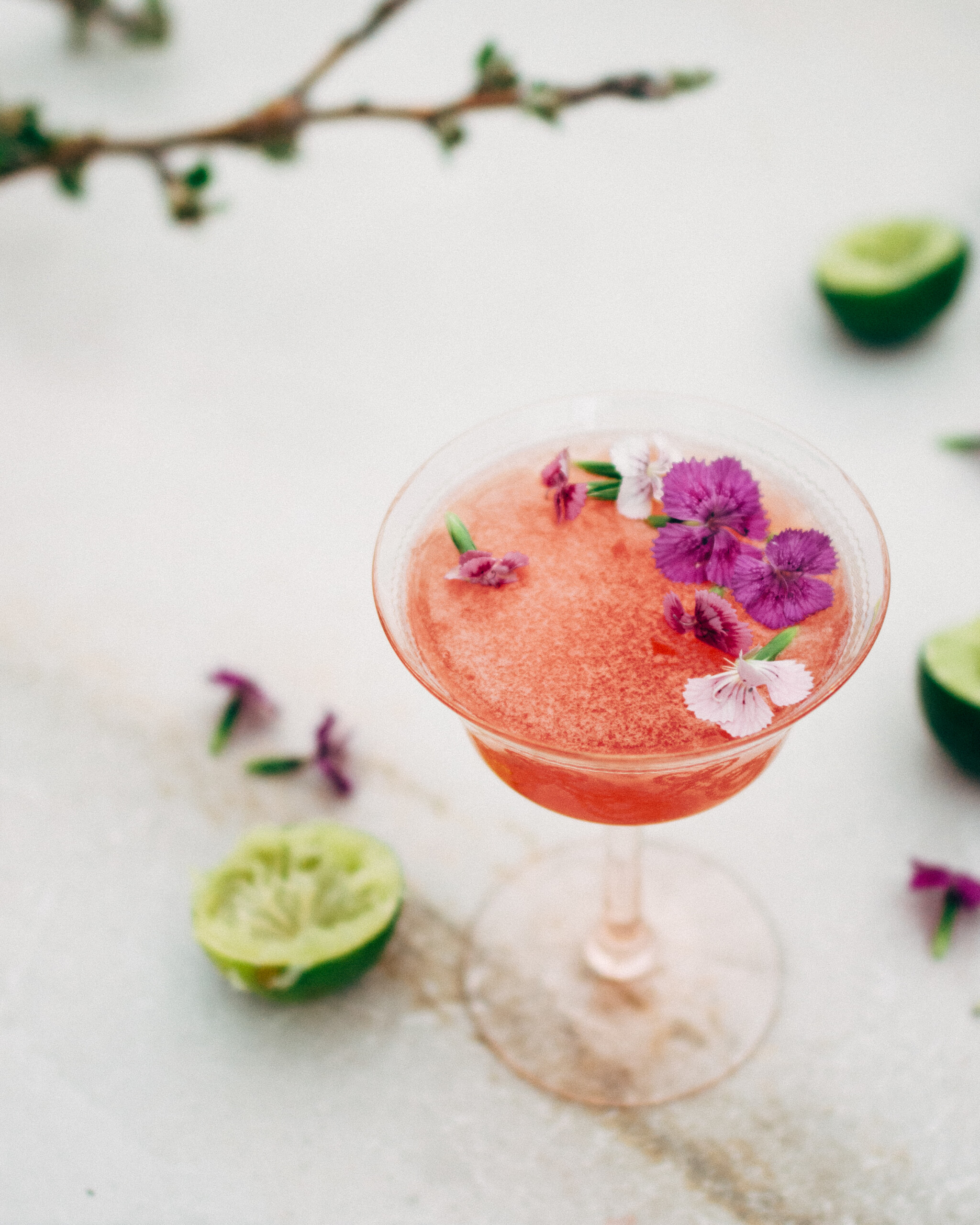 A side-view of a bright pink cocktail in a pale-pink cocktail glass, with edible purple flowers scattered on the drink's surface. There are freshly squeezed limes and scattered flowers in the background.