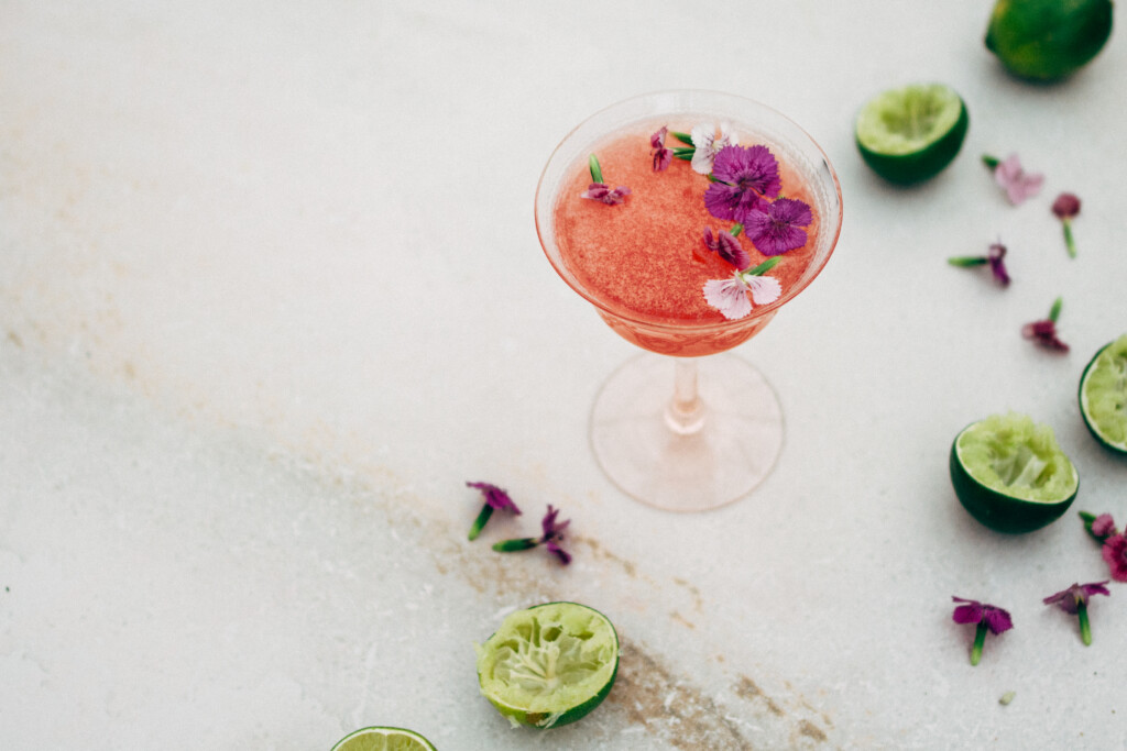 A side-view of a bright pink cocktail with edible purple flowers scattered on the drink's surface. There are freshly squeezed limes and scattered flowers in the background.